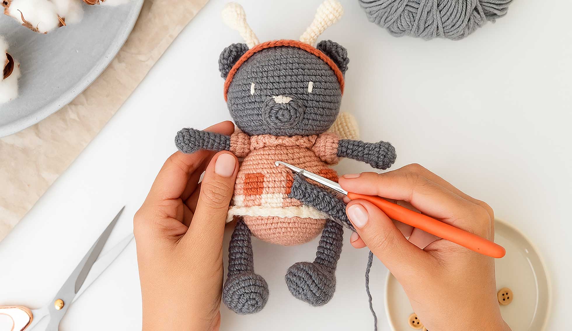 Manos tejiendo un amigurumi de oso con crochet, rodeado de hilo, botones y tijeras.